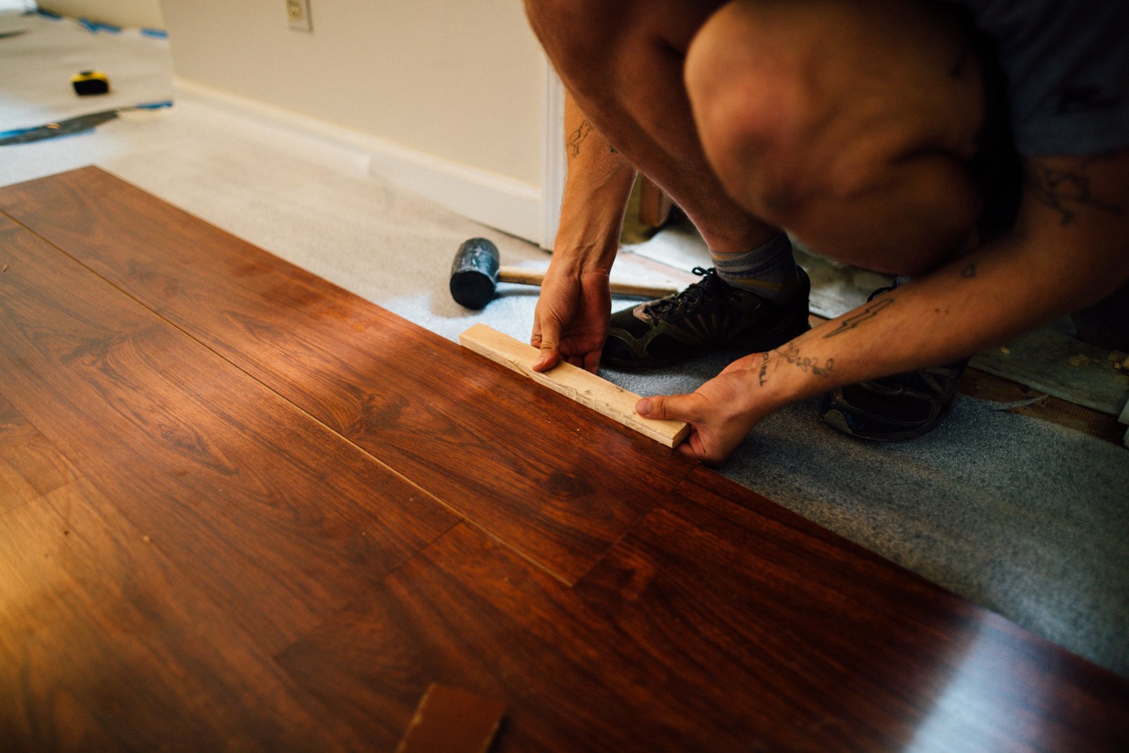 Hands tapping a laminate plank into place with a wood block, over grey underlay.
